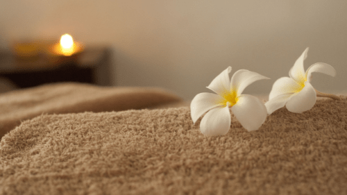 photo of a treatment be covered in a towell, with frangipani flowers on top and a candle glowing in the background