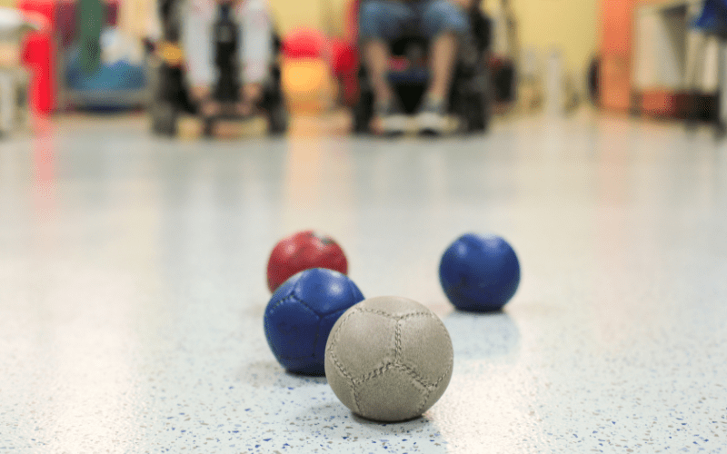 Photo of some different coloured balls on a smooth surface. People in the background are in wheelchairs.