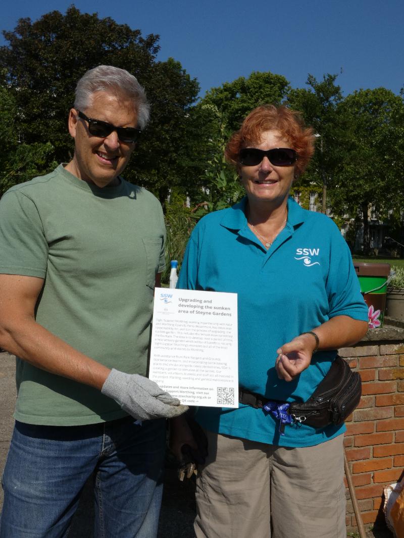 Photo of Cherry Ward, Chair of SSW's Board of Trustees, and volunteer Andrew, holding a piece of paper which explains the background of the sensory garden.