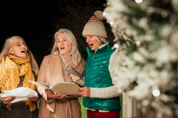 photo showing three women singing while stood outside next to a Christmas tree. The sky behind them is dark.