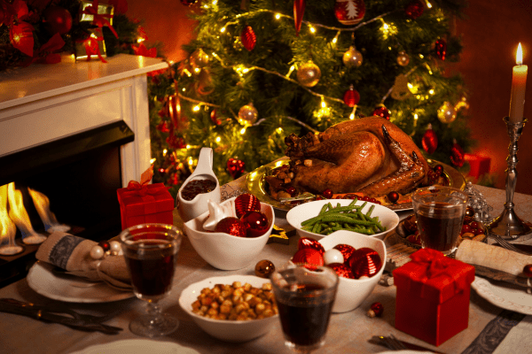 a photo of a Christma meal on a table, including turkey, vegetables, various sauces and glasses of red wine. There is a decorated Christmas tree in the background.