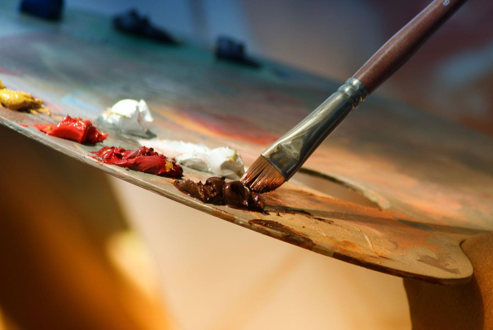 Paintbrush dipped in brown paint on wooden palette, with blobs of white and red paint,