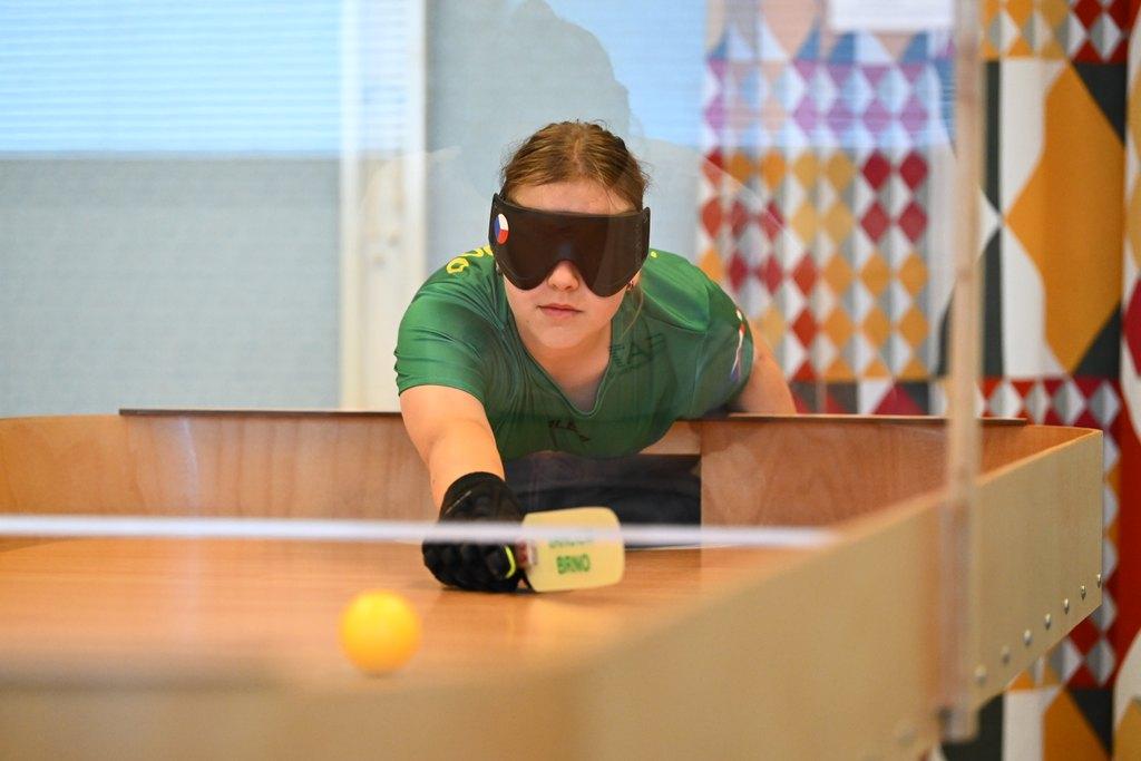 a photo of a young person playing Showdown. They are wearing a black eyemask and leaning forward over a large table. In their hand they have a small paddle, and there is a yellow ball on the table in front of them.