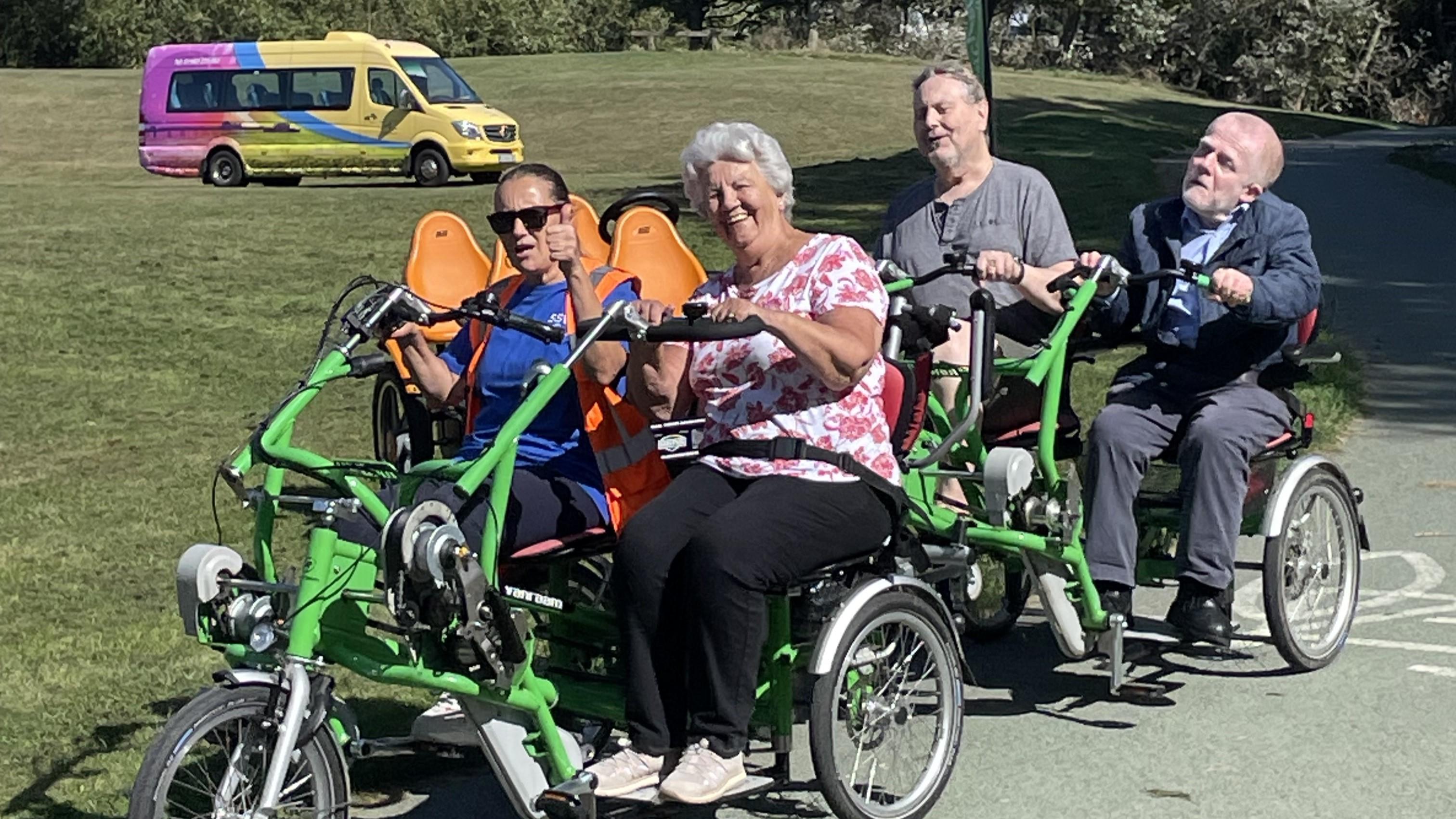 four ssw members riding on a four seater trike and trailer. The ssw minibus is parked on the field in the background
