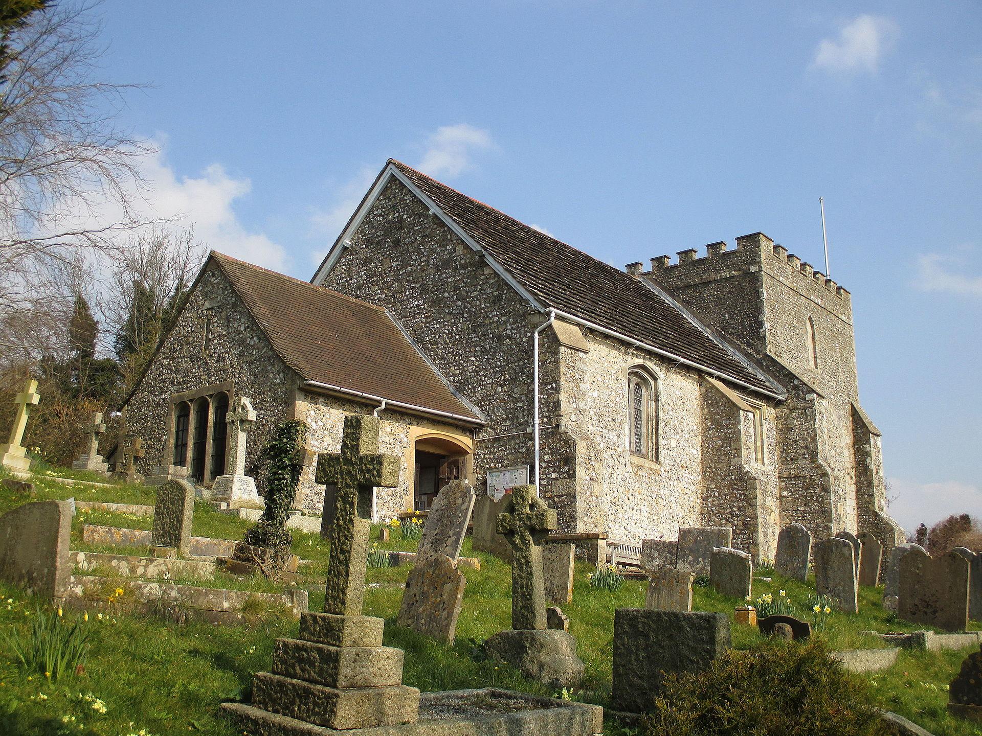 The photo is taken from the bottom of a grass slope and shows a stone church with a clear blue sky. in front of the church are an array of grave stones