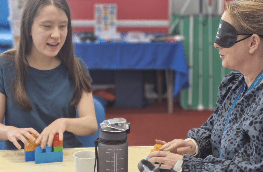 two people sitting together at a table. One has coloured blocks in front of them. The other is wearing a blind fold. They are talking to each other.