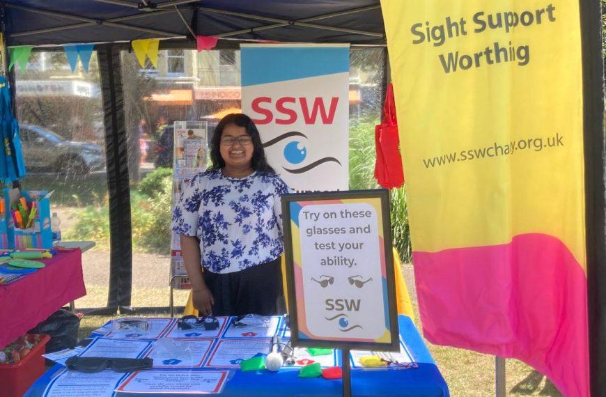 Poornima stands behind a table on the SSW stand. There are sim specs and leaflets on the table and a banner stand to the right.
