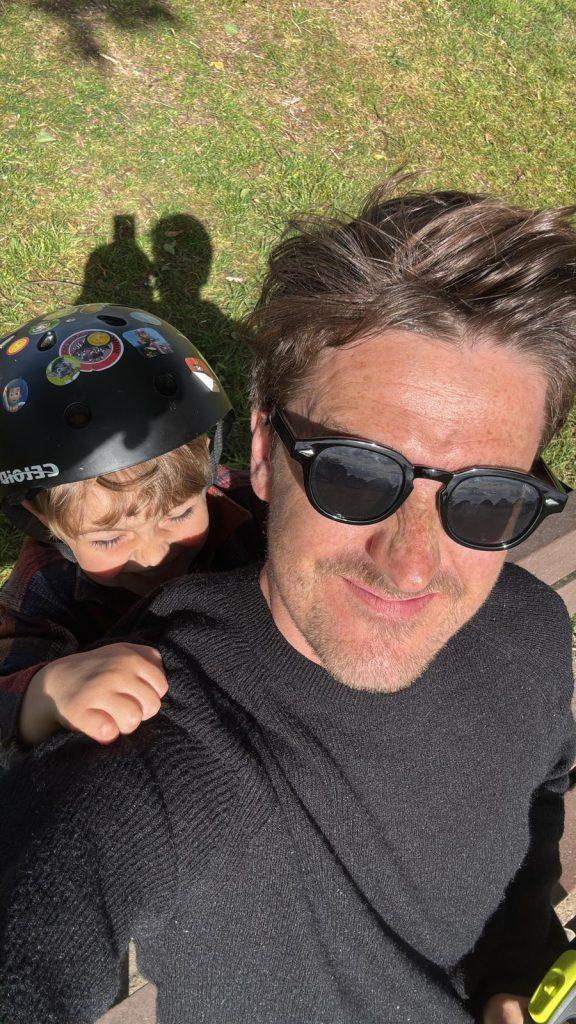 SSW LLHM team member Gary Playfoot lying on his back on grass, with his child.