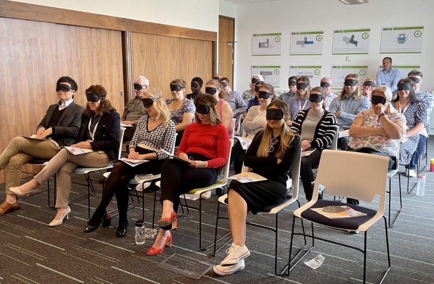 A room of delegates sat on rows of chairs in a conference room. The delegates are attending a VI awareness talk and are wearing blindfolds.