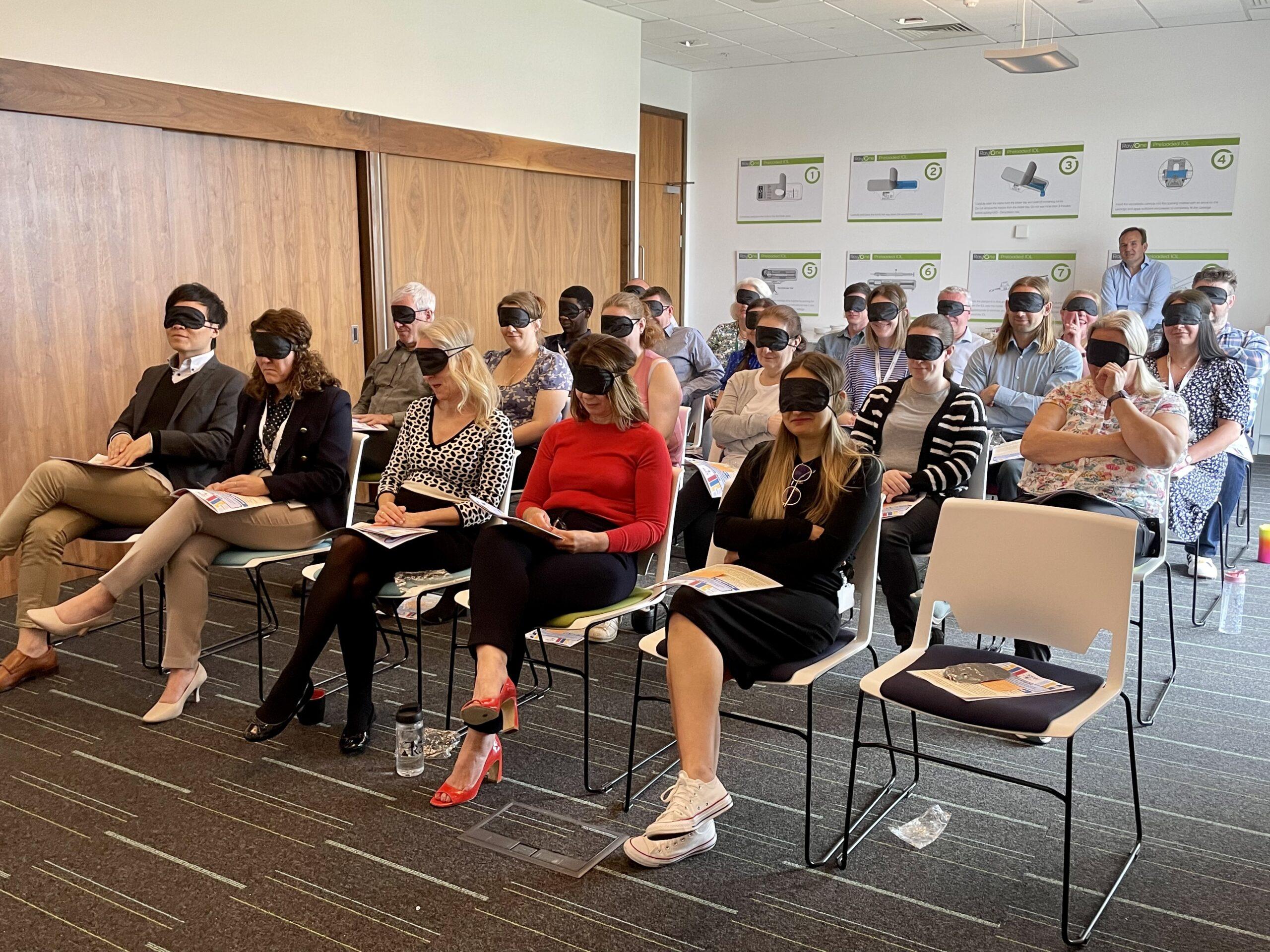 A room of delegates sat on rows of chairs in a conference room. The delegates are attending a VI awareness talk and are wearing blindfolds.
