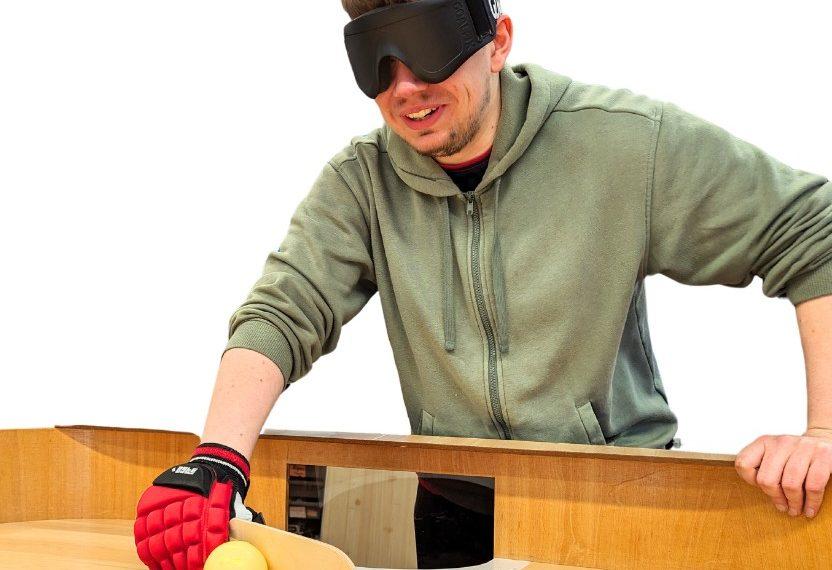 SSW client Sam playing Showdown. He is wearing a blindfol and holds a small paddle. There is a Showdown table in front of him, with a ball on it.